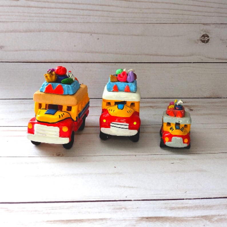 May include: Three colorful toy buses with red, yellow, and blue paint. The buses are decorated with fruit and vegetables on top. The buses are made of clay and are handmade.