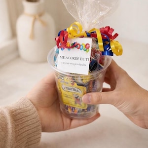 Op de afbeelding: Een doorzichtige plastic beker gevuld met verpakte snoepjes, bekroond met kleurrijke linten en een kleine kaart. De kaart leest "ME ACORDE DE TI" en "y te traje esta pendejadita". De beker wordt vastgehouden door een persoon die een beige trui draagt.