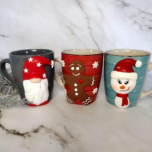 May include: Three ceramic Christmas mugs. One is gray with a gnome design, another is red with a gingerbread man, and the last is blue with a snowman. Each mug has a white handle and decorative elements.