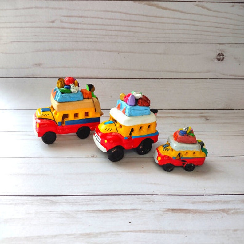 May include: Three colorful toy buses with yellow bodies, red wheels, and white roofs. The buses are loaded with colorful luggage and passengers. The buses are made of clay and are painted in a folk art style.