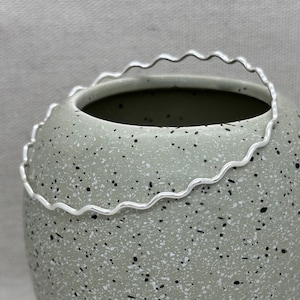 May include: A silver wavy bangle bracelet. The bracelet is sitting on a speckled green surface.