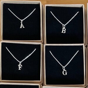 May include: A set of 18 silver necklaces with letter charms, displayed in individual boxes. Each box has a black interior and a beige exterior.