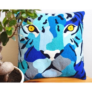 May include: Square cushion with a tiger face design in shades of blue, turquoise, and grey. The tiger's eyes are yellow, and the design is made with a textured, looped fabric. The cushion is placed on a wooden surface.