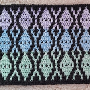 May include: A crocheted textile with a repeating diamond pattern. The diamonds transition in color from lavender to blue to green, outlined in black. The handmade item has a soft, textured appearance.