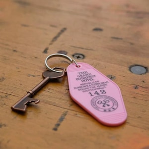 May include: Pink plastic hotel key chain with the text "The Grand Budapest Hotel" and "Republic of Zubrowka Nebelsbad Below the Alpine Sudetenwaltz" and the number "142".