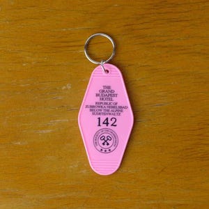 May include: Pink plastic hotel key chain with the text "The Grand Budapest Hotel, Republic of Zubrowka,  Nemelibad, Below the Alps,  Muertenwaltz, 142" and a logo of two crossed keys.