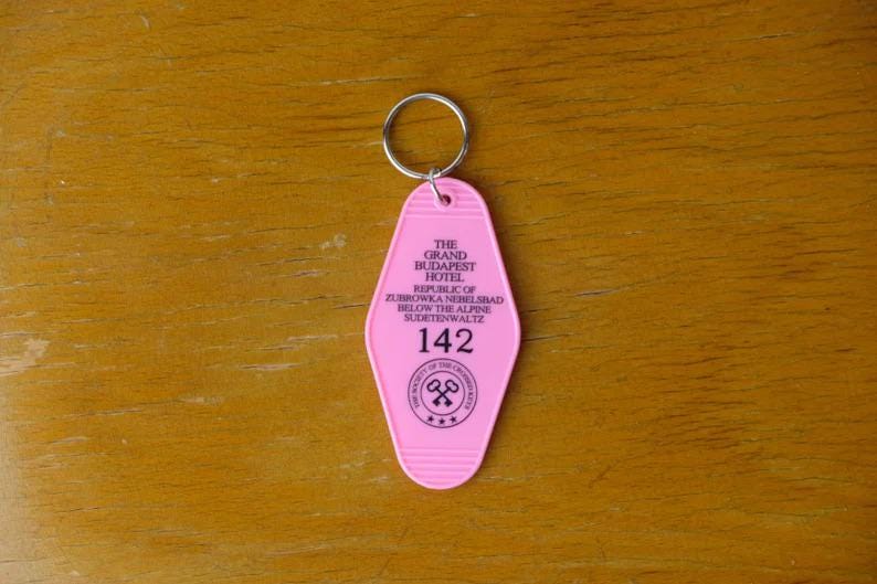 May include: Pink plastic hotel key chain with the text "The Grand Budapest Hotel, Republic of Zubrowka,  Nemelibad, Below the Alps,  Muertenwaltz, 142" and a logo of two crossed keys.