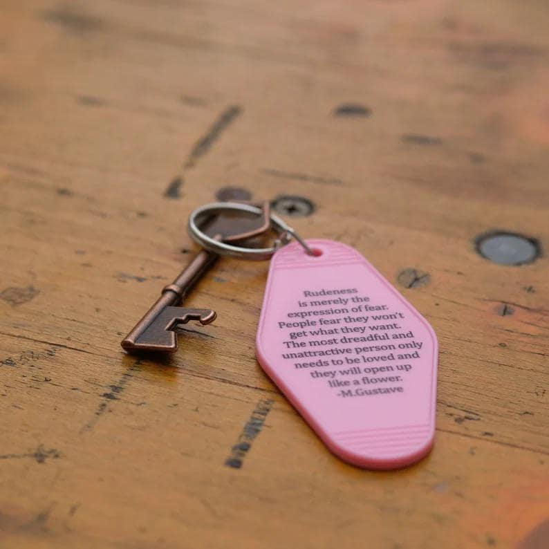 May include: A pink motel key tag with a quote about rudeness being an expression of fear. The quote is attributed to M. Gustave. The key tag is attached to a vintage-style skeleton key.
