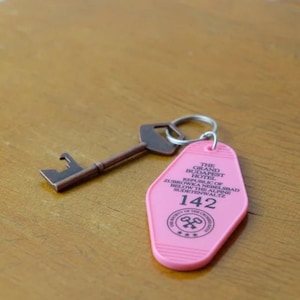 May include: Pink key card with black text for The Grand Budapest Hotel. The key card has the number 142 on it. A brown metal key is attached to the key card with a silver key ring.