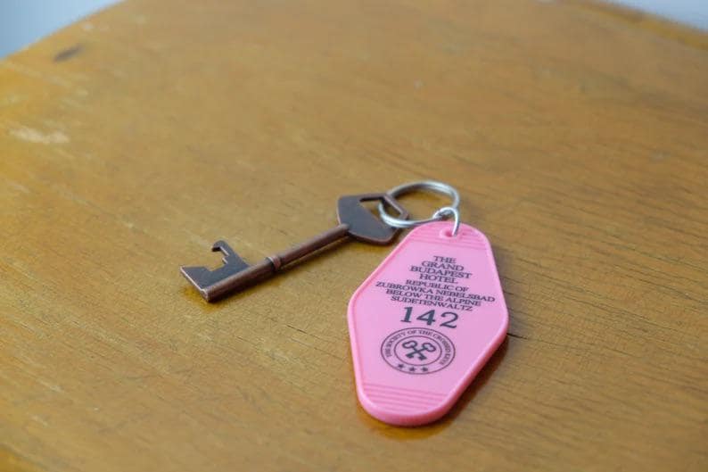 May include: Pink key card with black text for The Grand Budapest Hotel. The key card has the number 142 on it. A brown metal key is attached to the key card with a silver key ring.