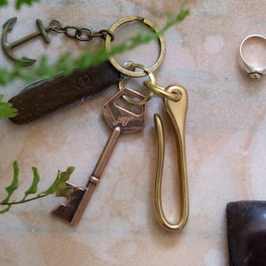 Op de afbeelding: Een sleutelhanger van messing met een sleutel, een haak en een ankerbedel. De sleutel heeft het woord "Joy" erop gegraveerd. De haak is een eenvoudige lus. De ankerbedel is een klein, gedetailleerd anker.