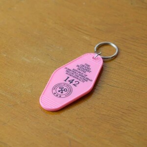 May include: Pink plastic key chain with a keyhole design. The keychain says "The Grand Budapest Hotel" and "Republic of Zubrowka" and "142".