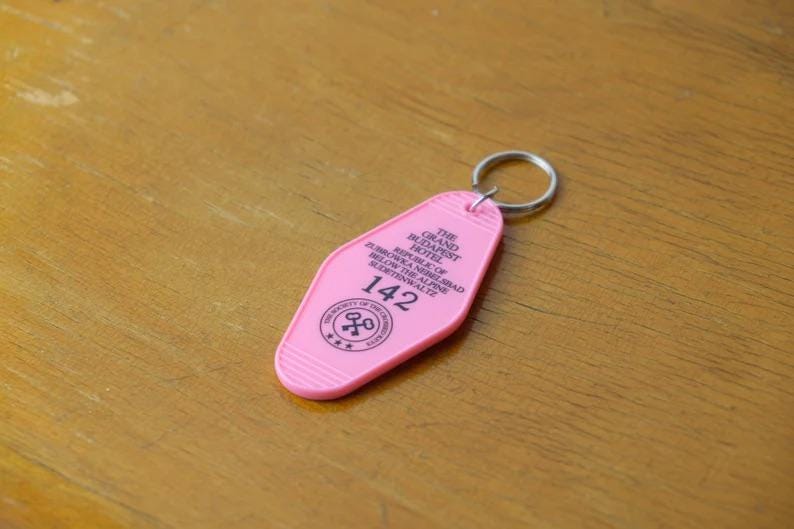 May include: Pink plastic key chain with a keyhole design. The keychain says "The Grand Budapest Hotel" and "Republic of Zubrowka" and "142".