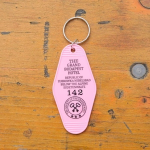 May include: Pink plastic hotel key chain with the text "The Grand Budapest Hotel" and "Republic of Zubrowka Nebelsbad Below the Alpine Sudetenwaltz" and the number "142". The key chain also has a logo with the text "The Society of the Crossed Keys" and three stars.