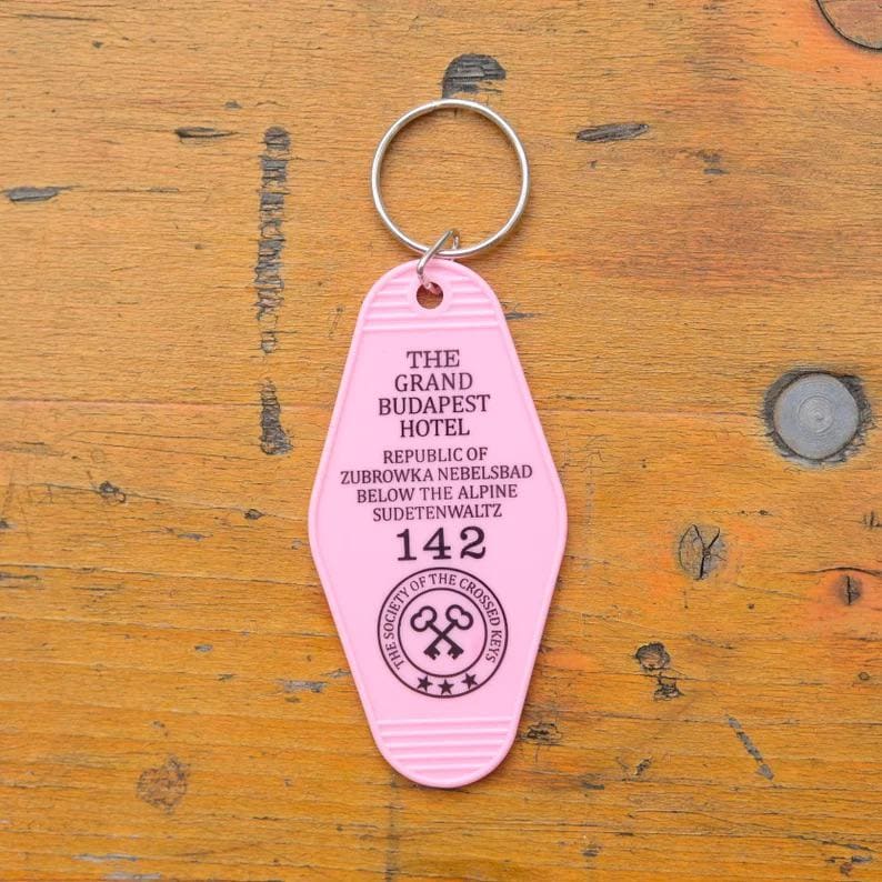 May include: Pink plastic hotel key chain with the text "The Grand Budapest Hotel" and "Republic of Zubrowka Nebelsbad Below the Alpine Sudetenwaltz" and the number "142". The key chain also has a logo with the text "The Society of the Crossed Keys" and three stars.