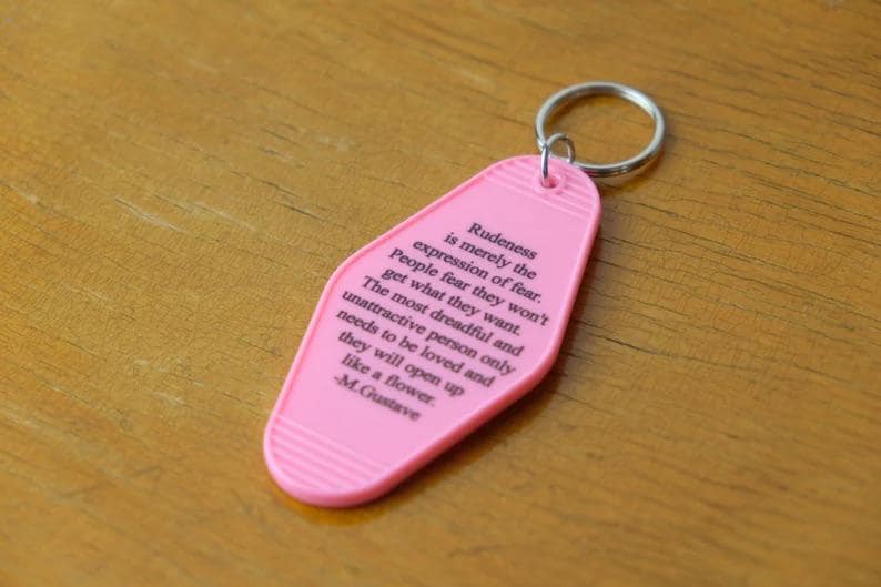 May include: Pink motel key chain with a quote about rudeness being an expression of fear. The quote is attributed to M. Gustave.