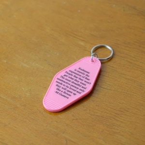 May include: Pink motel key chain with a quote about the expression of fear by M. Gustave. The quote reads: "Rakme is merely the expression of fear. People four they won't get what they want. The most dreadful and unattractive person only needs to be loved and they will open up like a flower. - M. Gustave"