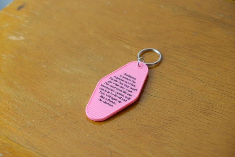May include: Pink motel key chain with a quote about the expression of fear by M. Gustave. The quote reads: "Rakme is merely the expression of fear. People four they won't get what they want. The most dreadful and unattractive person only needs to be loved and they will open up like a flower. - M. Gustave"