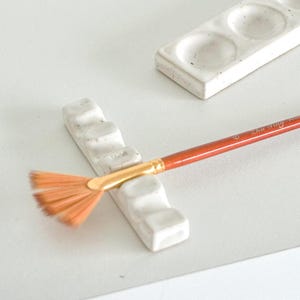 Op de afbeelding: Een keramische kunstenaarspalet en penseelhouder set. De set bevat een rechthoekig palet met drie putjes en een penseelhouder met een sleuf voor een penseel. Een oranje handvat waaierpenseel rust in de houder.