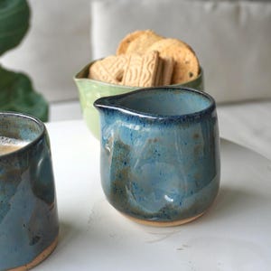 Könnte beinhalten: Eine blaue Keramik-Sahnekännchen mit Ausguss, mit gesprenkelter Glasur. Daneben eine passende Tasse und eine grüne Schüssel mit Keksen. Die Keramik hat eine rustikale, handgemachte Ästhetik, ideal für Kaffee oder Tee.