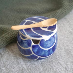 Może przedstawiać: Sólniczka ceramiczna w kolorze niebieskim i białym z dopasowaną drewnianą łyżeczką. Sólniczka ma falisty wzór i pokrywkę z małym otworem na łyżeczkę.