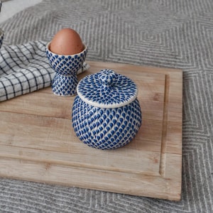 May include: A blue and white ceramic jar with a lid, and a blue and white ceramic egg cup with an egg inside. The jar and egg cup are on a wooden chopping board.