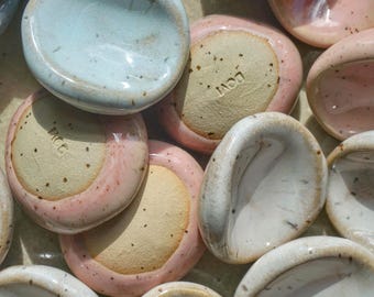 May include: A collection of small, ceramic dishes in various pastel colors, including pink, blue, and white. The dishes have a smooth, glossy finish and a shallow, rounded shape. The underside of some dishes has a stamped text.