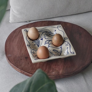 May include: A square, cream-coloured ceramic egg holder with a blue illustrated design, holding three brown eggs. The holder sits on a dark brown wooden tray, with a grey cushion in the background.