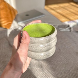 Puede incluir: Primer plano de una mano sosteniendo una taza de cerámica gris claro con una bebida de matcha verde en su interior. La taza tiene una superficie texturizada y está apilada en tres capas.