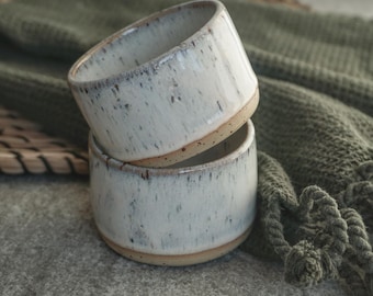 May include: Two white ceramic bowls with a speckled glaze. The bowls are stacked on top of each other and have a light brown rim.