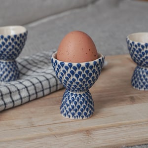 Può includere: Due porta uova in ceramica blu e bianca con un motivo blu e bianco. Un porta uova contiene un uovo marrone.