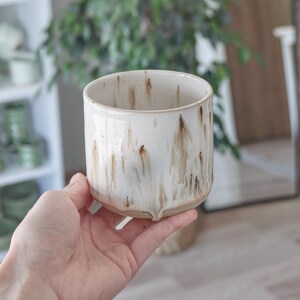 May include: A cylindrical ceramic cup featuring a glossy off-white glaze with vertical brown drip patterns. The base of the cup is unglazed, revealing a natural tan clay texture. The rim is smooth and circular, with a consistent wall thickness.