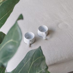 Puede incluir: Dos tazas pequeñas de cerámica blanca con asas sobre una superficie texturizada de color beige claro. Las tazas tienen un diseño simple y minimalista. Hojas verdes enmarcan las tazas en primer plano.