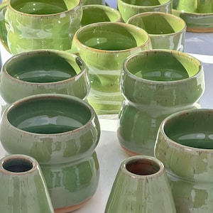 Op de afbeelding: Een verzameling handgemaakte groene keramiek, waaronder mokken, kommen en een kan. De stukken hebben een glanzende afwerking en een rustieke uitstraling. De collectie omvat mokken met handvatten, kleine kommen en een grotere kan.