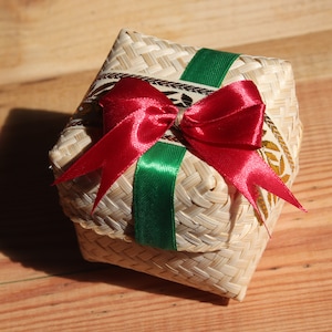 May include: A small, woven basket with a red satin ribbon tied in a bow. The basket is decorated with a green ribbon and a gold pattern.