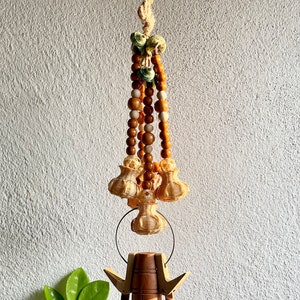 May include: A wooden wind chime with a brown, cylindrical base and three round, wooden bells. The chime is suspended from a string of brown and white beads with green and brown accents.
