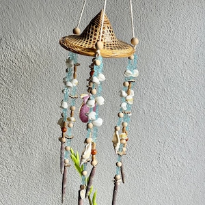 Op de afbeelding: Een geweven bamboehoed met een kralenmobiel dat eraan hangt. Het mobiel heeft blauwe glazen kralen, witte schelpen en houten kralen. Het mobiel hangt aan een wit touw.