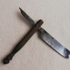 Könnte beinhalten: Ein antikes Klappmesser mit einem dunklen Holzgriff und einer angelaufenen Stahlklinge. Die Klinge ist geöffnet und zeigt ihre volle Länge. Das Messer scheint abgenutzt zu sein, was auf Alter und Gebrauch hindeutet. Der Griff hat ein abgerundetes Ende.