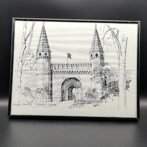 Op de afbeelding: Zwart omlijnde pen- en inkttekening van het Topkapi-paleis in Istanbul, Turkije. Het kunstwerk toont de ingang van het paleis met twee hoge torens, een gewelfde poort en een kleine vlag. De tekst "TOPKAPI PALACE - ISTANBUL - 5-26-96" staat onderaan.