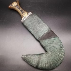 May include: An antique dagger with a curved blade and a decorative handle. The handle is gold-toned with circular embellishments, and the blade is wrapped in green cord. The dagger has a dark, aged appearance.