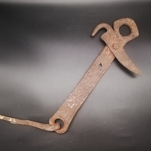 May include: A rusty, antique metal tool with a hook, a pointed blade, and a patterned handle. The tool has a loop at the end of the handle and is connected to a thin, flat metal piece. The tool is likely used for a specific task.