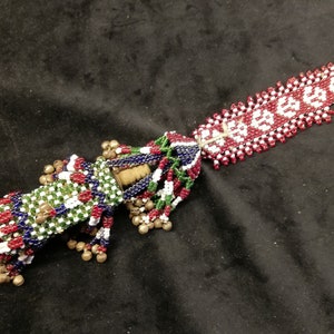 Puede incluir: Un objeto decorativo con cuentas con un mango rectangular y una borla. El mango es verde, blanco, rojo y azul. El mango tiene pequeñas campanas doradas. El mango rectangular es rojo y blanco con un estampado floral.