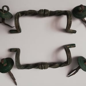May include: An antique bronze hardware set, comprising two twisted-design handles and four rosette-shaped decorative elements. The handles have a curved design, and the rosettes are circular with a central loop. The set has a weathered, aged appearance.