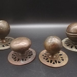 May include: Four antique bronze door knobs with decorative circular bases. The knobs have various spherical and oval shapes. The bases feature intricate cut-out designs. The knobs are displayed against a dark background.
