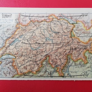 以下が含まれることがあります： スイスのヴィンテージ長方形地図。詳細な地形、政治的境界線、アラビア文字で書かれた地理的ラベルが特徴で、薄い青色の枠線が付いたクリーム色の紙に印刷されています。
