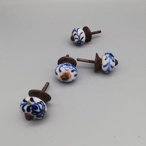 May include: Four ceramic cabinet knobs with a white base and blue floral design. Each knob has a rusty brown metal base and screw. The knobs are a decorative hardware element for furniture.