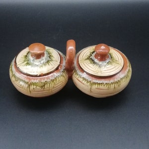 May include: A set of two small ceramic containers with lids. The containers are brown with green and white painted designs. The containers are connected by a handle.
