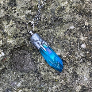 May include: A silver chain necklace with a blue crystal pendant. The pendant is shaped like a long, pointed crystal with a silver cap at the top.