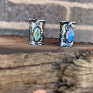 May include: Two silver-coloured, cylindrical rings with textured surfaces. One ring features a green and yellow gemstone, the other a blue gemstone. The rings are displayed on a wooden surface, with a brick background.