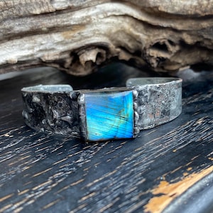 Puede incluir: Un brazalete de plata con una piedra preciosa de labradorita azul iridiscente cuadrada engastada en el centro. El brazalete tiene un acabado texturizado y martillado.